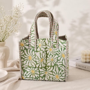 May include: A quilted tote bag featuring a repeating pattern of white daisy flowers with yellow centers on a green background. The bag has two sturdy handles with a red, white, and green striped trim that matches the edges of the square-shaped bag.