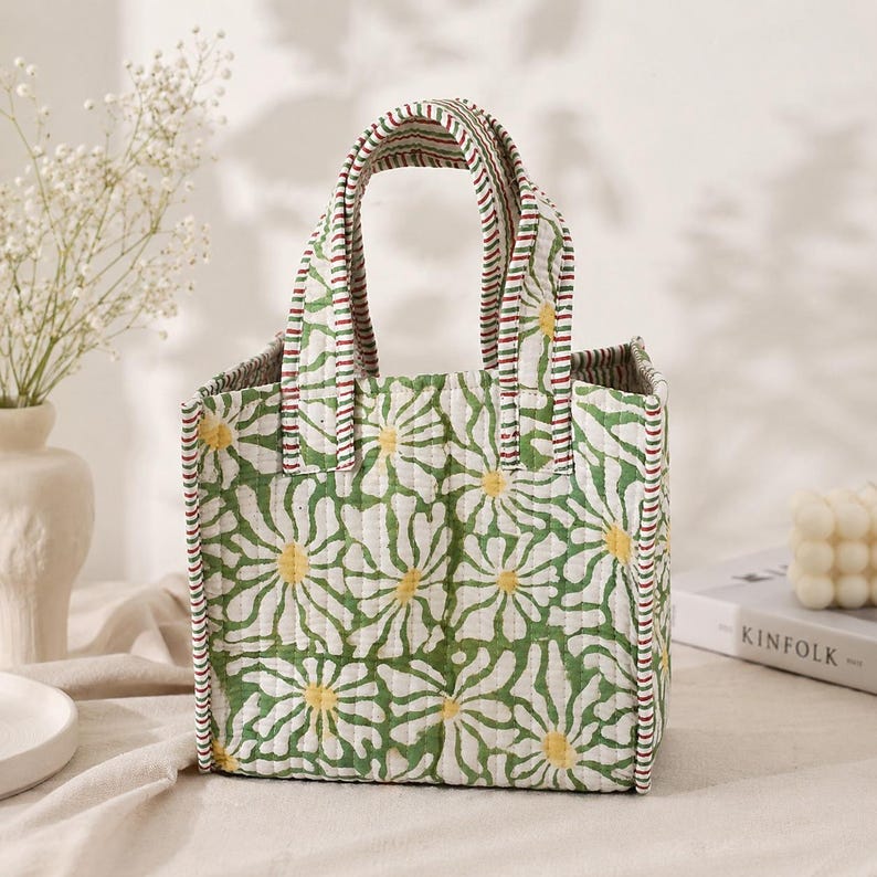 May include: A quilted tote bag featuring a repeating pattern of white daisy flowers with yellow centers on a green background. The bag has two sturdy handles with a red, white, and green striped trim that matches the edges of the square-shaped bag.