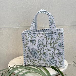 May include: A quilted tote bag featuring a white base with a green and blue floral pattern. The bag has two top handles and side panels decorated with thin horizontal green and white stripes, providing a structured, rectangular silhouette.