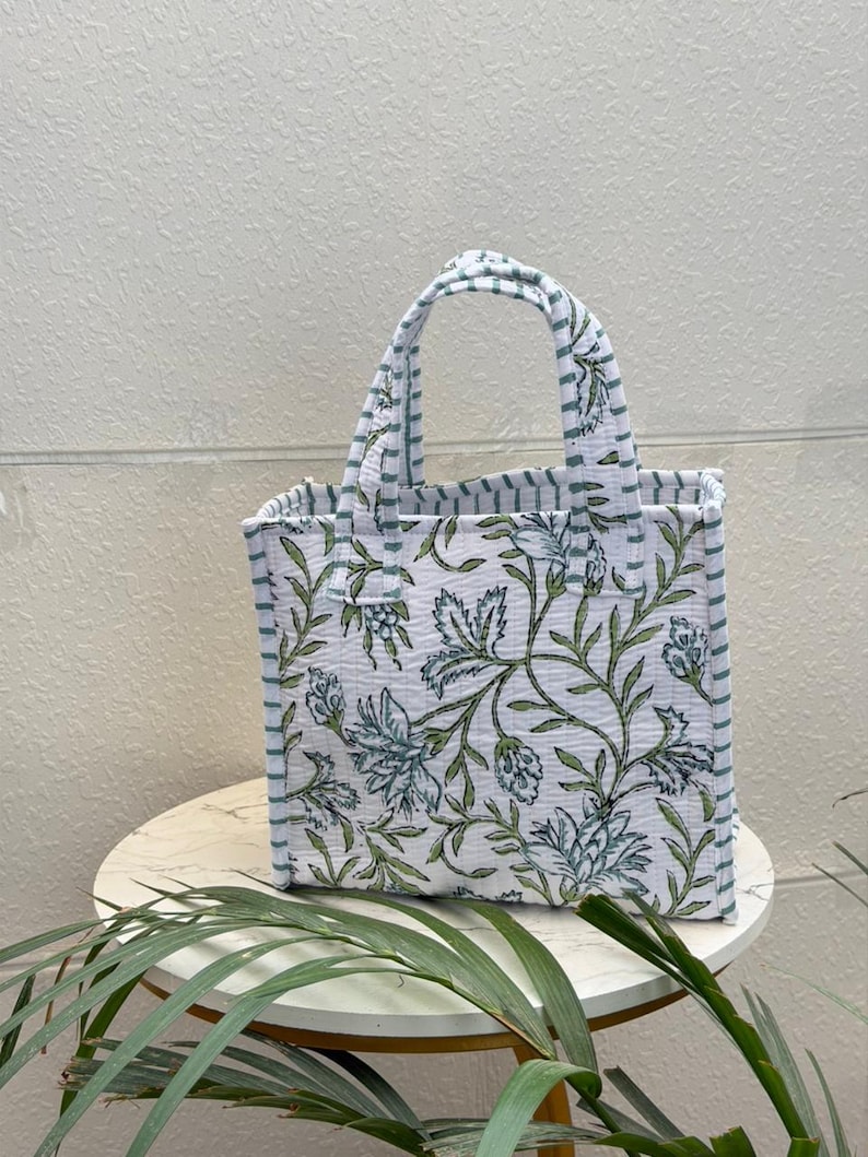 May include: A quilted tote bag featuring a white base with a green and blue floral pattern. The bag has two top handles and side panels decorated with thin horizontal green and white stripes, providing a structured, rectangular silhouette.