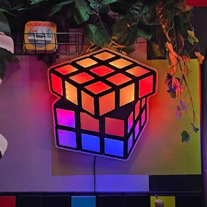 Puede incluir: Una lámpara de estilo Rubik's Cube, multicolor y brillante, con un marco negro. La lámpara está montada en una pared con un fondo de azulejos blanco y negro. La hora 23:38 se muestra en un reloj digital en el fondo.
