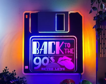 Pode incluir: Uma placa de néon em forma de disquete com o texto "Back to the 90's Retro Love" em letras brancas. A placa é iluminada em um arco-íris de cores, incluindo vermelho, laranja, amarelo, verde, azul e roxo. Dois crânios de açúcar coloridos estão em primeiro plano.