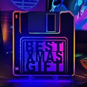 Könnte beinhalten: Ein schwarzes und weißes 3,5 Zoll Floppy-Disk-förmiges Licht mit einem regenbogenfarbenen Rand. Das Licht hat den Text "BEST XMAS GIFT" im Inneren.