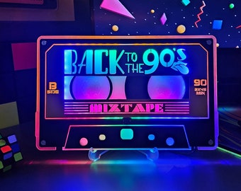 Pode incluir: Uma luz preta em forma de cassete com luzes LED coloridas. A luz apresenta o texto "Back to the 90's Mixtape" e "90 2X45 MIN".