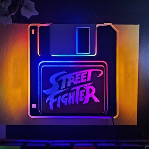 May include: A neon sign shaped like a floppy disk with the words "Street Fighter" in white lettering on a blue and pink background.
