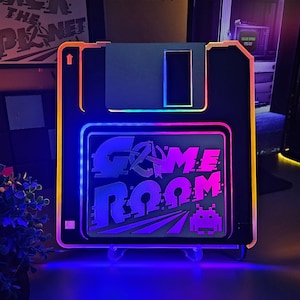 May include: Black, retro-style Game Room sign in the shape of a floppy disk, with a colorful LED border. The sign features the words "GAME ROOM" in a pixelated font, and an image of an arcade game.