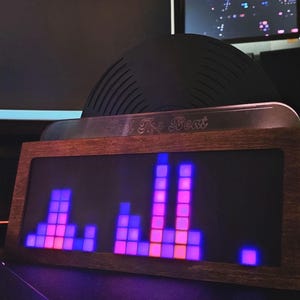 May include: A rectangular wooden audio visualiser featuring a dark brown frame and a digital display with glowing purple and pink square bars. The device has a metallic top plate with the engraved text "Feel The Beat" and three circular control knobs.
