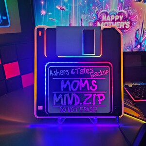 May include: A black and white floppy disk shaped light with a blue, pink and purple glow. The disk has the text "Asher's & Tate's backup MOMS MIND.ZIP DO NOT ERASE!" on it.