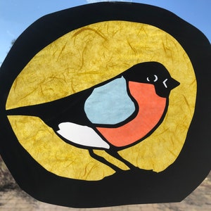 May include: A colourful paper craft of a bird with a black body, white chest, red belly, and blue wings. The bird is set against a yellow background with a black circle around it.
