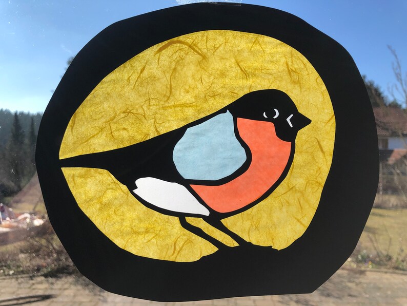 May include: A colourful paper craft of a bird with a black body, white chest, red belly, and blue wings. The bird is set against a yellow background with a black circle around it.