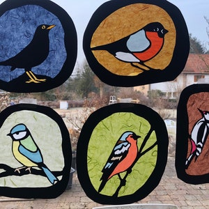 May include: Five colourful stained glass circles with black outlines, each featuring a different bird. The birds are a blackbird, a robin, a blue tit, a goldfinch, and a woodpecker.