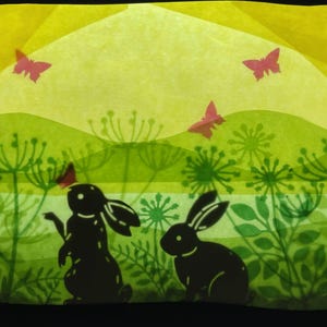May include: A charming illustration of two black rabbit silhouettes in a green meadow, with pink butterflies against a yellow and green backdrop. The artwork depicts a springtime scene, with stylized plants and a whimsical feel.