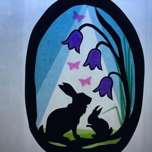 May include: A black silhouette of two rabbits beneath purple bell-shaped flowers and pink butterflies, framed in an oval. The background has blue and white stripes, resembling a sky.