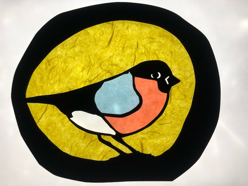 May include: A colourful paper cutout of a bird with a black body, white chest, blue wing, and red breast. The bird is set against a yellow background inside a black circle.
