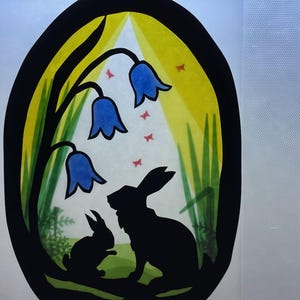 May include: An oval artwork depicting two rabbit silhouettes, bluebell flowers, and red butterflies. The background is yellow and green, framed by a black border. The image has a whimsical, nature-inspired theme, reminiscent of a stained-glass window.