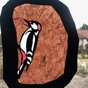 Pode incluir: Decora&ccedil;&atilde;o de janela com um desenho de pica-pau. A ave &eacute; ilustrada em preto, branco e rosa sobre um fundo castanho texturizado, emoldurado por uma borda preta. A obra de arte est&aacute; colocada em frente a uma cena externa desfocada, vis&iacute;vel atrav&eacute;s da janela.