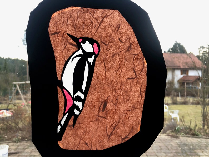 Pode incluir: Decora&ccedil;&atilde;o de janela com um desenho de pica-pau. A ave &eacute; ilustrada em preto, branco e rosa sobre um fundo castanho texturizado, emoldurado por uma borda preta. A obra de arte est&aacute; colocada em frente a uma cena externa desfocada, vis&iacute;vel atrav&eacute;s da janela.