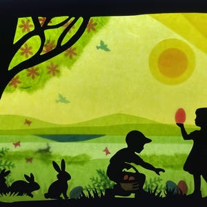 May include: Silhouette artwork depicting an Easter scene with a tree, rabbits, and children. A boy and girl are collecting eggs, with one holding a red egg. The background shows a sun, birds, and rolling hills, in green and yellow hues.