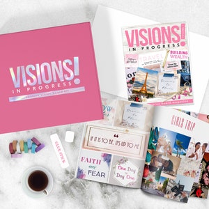 May include: Pink box with the text "VISIONS! IN PROGRESS" and the words "couture vision board kit".  A magazine with the text "VISIONS! IN PROGRESS" and the words "vision board magazine".  A magazine open to a page with the text "GIRLS TRIP" and images of a beach, a passport, and a city.  A glue stick, a cup of coffee, and a stack of colorful washi tape.