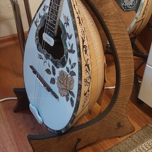 May include: A white and brown bouzouki with intricate floral inlays and a black leather strap is displayed on a wooden stand. The stand has a black leather strap and a small logo on the bottom.