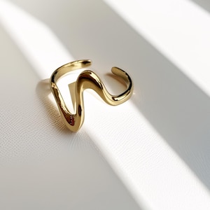 Könnte beinhalten: Ein goldfarbener Ring mit wellenförmigem Design. Der Ring ist unten offen und hat eine glatte, polierte Oberfläche. Der Ring befindet sich vor einem weißen Hintergrund mit Schatten.