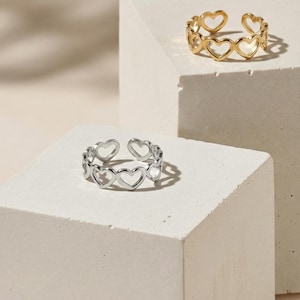 Könnte beinhalten: Zwei offene Ringe, einer goldfarben und einer silberfarben, jeweils mit herzförmigen Aussparungen. Die Ringe werden auf weißen geometrischen Blöcken vor neutralem Hintergrund präsentiert und zeigen ein romantisches Schmuckdesign.