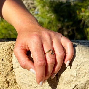 Könnte beinhalten: Ein goldfarbener Ring mit einem durchgehenden, wellenförmigen Design, das sich um den Finger legt. Das Band hat eine glatte, polierte metallische Oberfläche, die das Licht reflektiert und diesem Schmuckstück eine schlichte Optik verleiht.