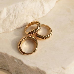 Könnte beinhalten: Drei goldfarbene Ringe mit einem strukturierten, seilartigen Design. Die Ringe werden auf einer hellen, strukturierten Oberfläche präsentiert. Die Ringe sind ein schlichtes, aber elegantes Schmuckstück.