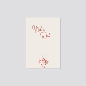 Könnte beinhalten: Eine cremefarbene Karte mit den Worten "Make a Wish" in roter Schrift. Unter dem Text befinden sich drei rote Sternschnuppen. Die Karte ist rechteckig und vor einem hellgrauen Hintergrund platziert.