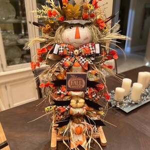 Peut inclure: Une d&eacute;coration d'automne sur le th&egrave;me de l'&eacute;pouvantail avec un support en bois, comprenant un visage d'&eacute;pouvantail, un tissu &agrave; th&egrave;me automnal, des citrouilles et une pancarte qui dit "Je pense que j'aime l'automne plus que tout".