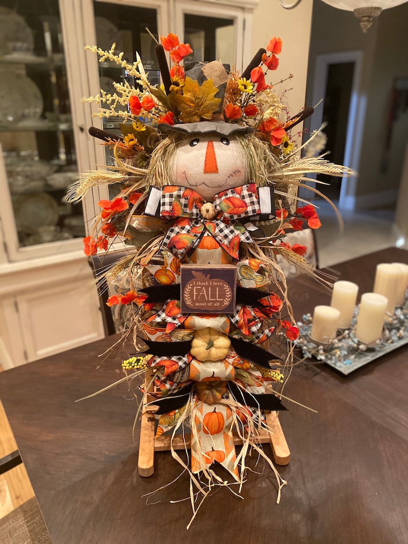 Peut inclure: Une d&eacute;coration d'automne sur le th&egrave;me de l'&eacute;pouvantail avec un support en bois, comprenant un visage d'&eacute;pouvantail, un tissu &agrave; th&egrave;me automnal, des citrouilles et une pancarte qui dit "Je pense que j'aime l'automne plus que tout".