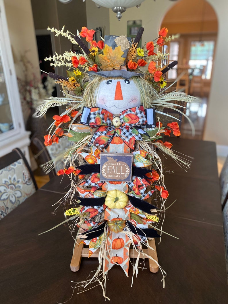 Peut inclure: Une d&eacute;coration de porte d'automne sur le th&egrave;me de l'&eacute;pouvantail avec une base en toile de jute et en paille, un visage en tissu avec un n&oelig;ud et une pancarte qui dit "I think I love FALL most of all".