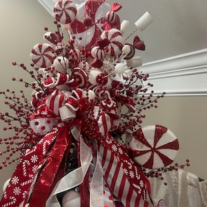 May include: A festive Christmas tree topper featuring red and white candy-themed ornaments. The arrangement includes large peppermint swirl decorations, candy canes, and red berry sprigs, all accented with red and white ribbons and bows.