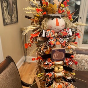 Peut inclure: Une d&eacute;coration de porte d'automne sur le th&egrave;me de l'&eacute;pouvantail avec une base en bois, une t&ecirc;te d'&eacute;pouvantail envelopp&eacute;e de toile de jute et une pancarte qui dit "Je pense que j'aime l'automne plus que tout". La d&eacute;coration est orn&eacute;e de rubans aux couleurs d'automne, de citrouilles et d'accents floraux.