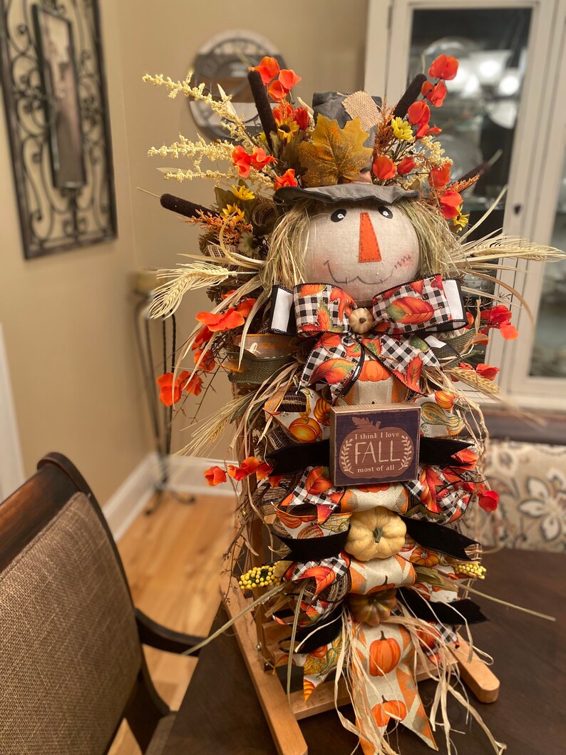 Peut inclure: Une d&eacute;coration de porte d'automne sur le th&egrave;me de l'&eacute;pouvantail avec une base en bois, une t&ecirc;te d'&eacute;pouvantail envelopp&eacute;e de toile de jute et une pancarte qui dit "Je pense que j'aime l'automne plus que tout". La d&eacute;coration est orn&eacute;e de rubans aux couleurs d'automne, de citrouilles et d'accents floraux.