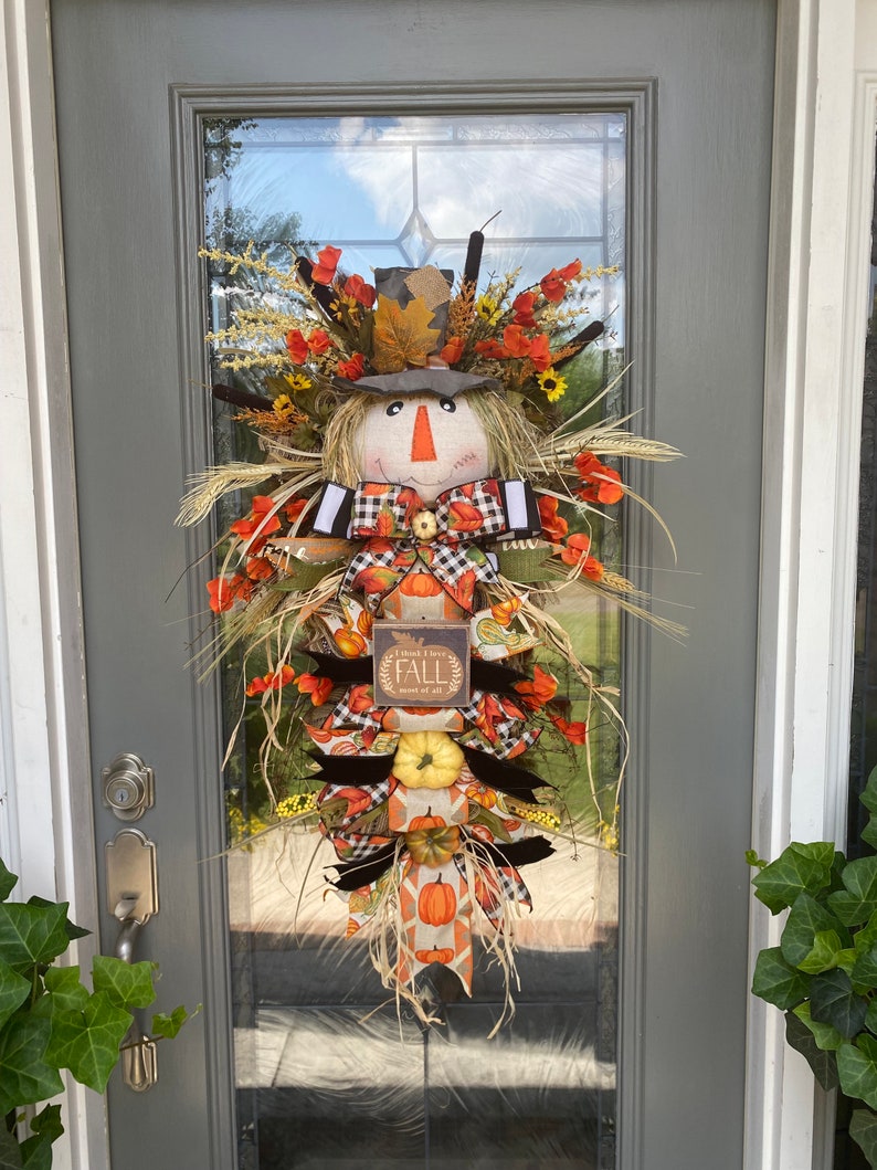 Peut inclure: Une couronne de porte sur le th&egrave;me de l'automne avec un visage de  &eacute;pouvantail, des citrouilles et une pancarte qui dit "Je pense que j'aime l'automne plus que tout".