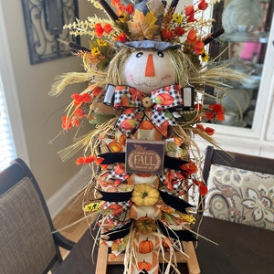 Peut inclure: Une d&eacute;coration de scarecrow sur le th&egrave;me de l'automne avec un visage en toile de jute, un chapeau bleu et un n&oelig;ud en tissu &agrave; carreaux. Le scarecrow est entour&eacute; de feuillage d'automne, y compris des tiges de bl&eacute;, des citrouilles et des fleurs orange. Une pancarte en bois avec le texte "Je pense que j'aime l'automne plus que tout" est attach&eacute;e au scarecrow.