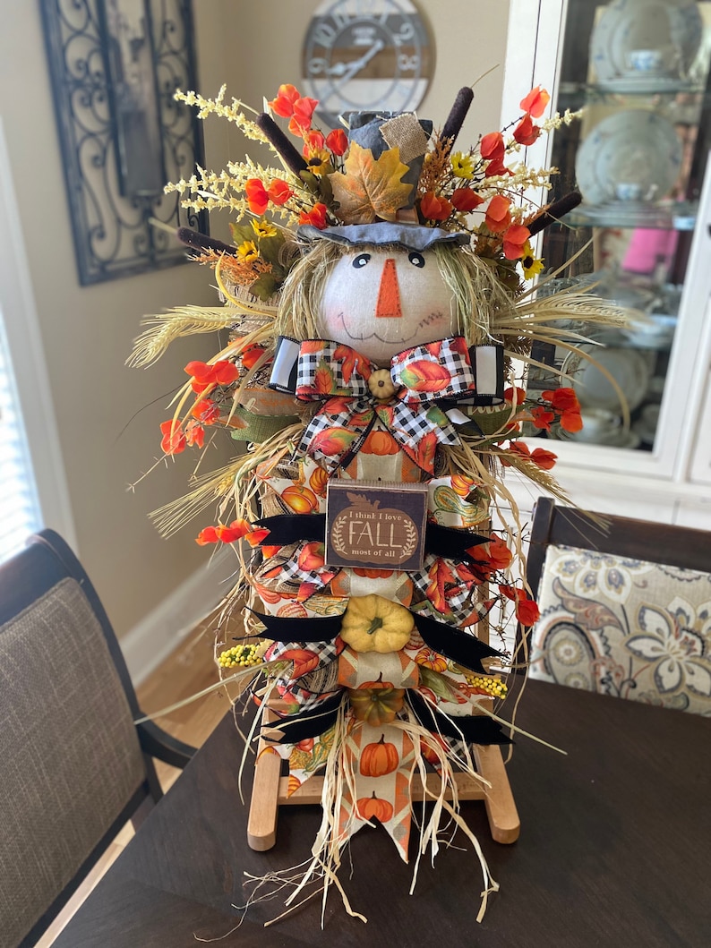 Peut inclure: Une d&eacute;coration de scarecrow sur le th&egrave;me de l'automne avec un visage en toile de jute, un chapeau bleu et un n&oelig;ud en tissu &agrave; carreaux. Le scarecrow est entour&eacute; de feuillage d'automne, y compris des tiges de bl&eacute;, des citrouilles et des fleurs orange. Une pancarte en bois avec le texte "Je pense que j'aime l'automne plus que tout" est attach&eacute;e au scarecrow.