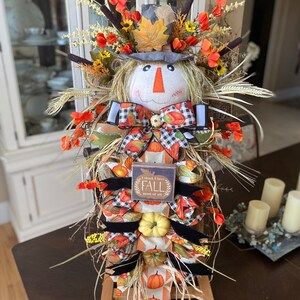 Peut inclure: Une d&eacute;coration de porte d'automne sur le th&egrave;me de l'&eacute;pouvantail avec une base en toile de jute, un visage d'&eacute;pouvantail, un n&oelig;ud &agrave; carreaux noir et blanc et une pancarte qui dit "Je pense que j'aime l'automne plus que tout".