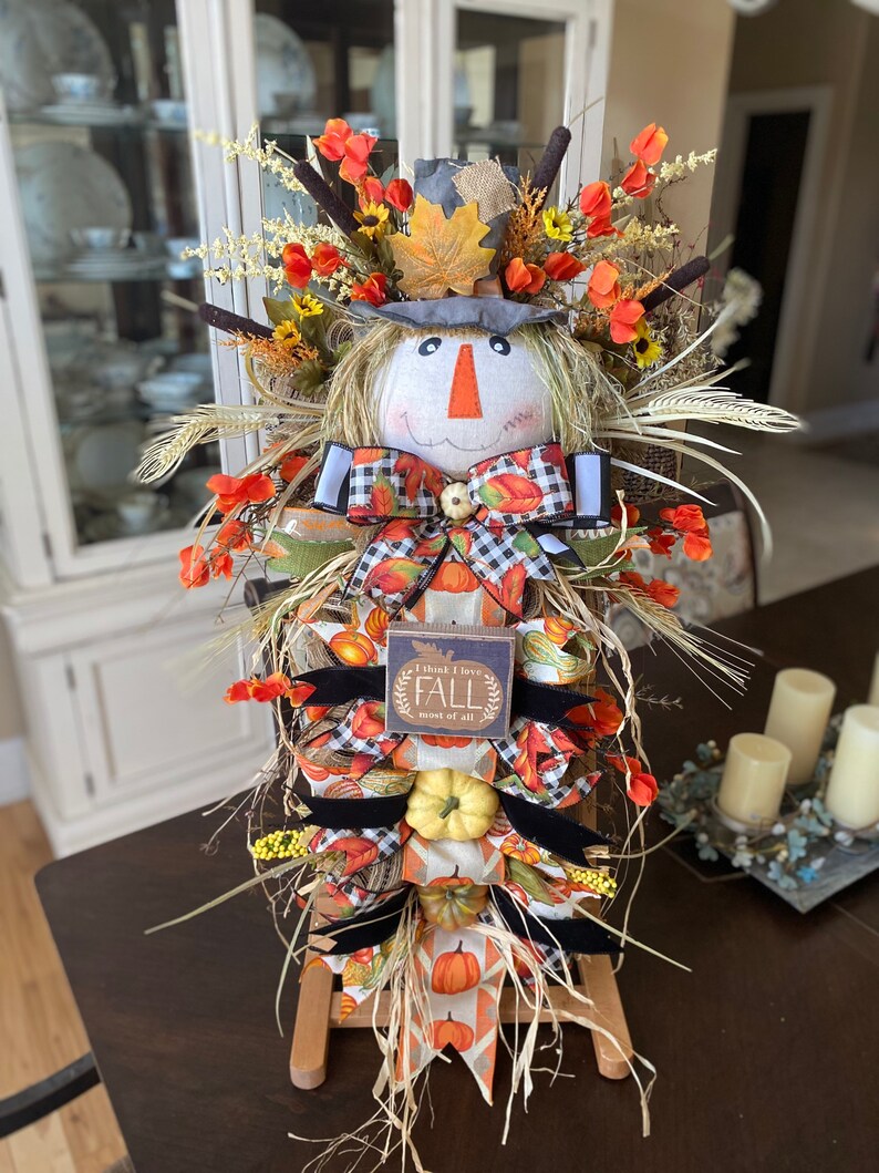 Peut inclure: Une d&eacute;coration de porte d'automne sur le th&egrave;me de l'&eacute;pouvantail avec une base en toile de jute, un visage d'&eacute;pouvantail, un n&oelig;ud &agrave; carreaux noir et blanc et une pancarte qui dit "Je pense que j'aime l'automne plus que tout".