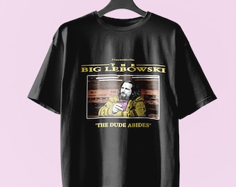 Könnte beinhalten: Ein schwarzes Kurzarm-T-Shirt mit einem Grafikdruck eines Mannes in einem gelben gemusterten Pullover, der ein Glas hält. Der Text A Coen Brothers Film, THE BIG LEBOWSKI und THE DUDE ABIDES ist in gelben und weißen Buchstaben aufgedruckt.
