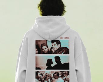 Könnte beinhalten: Ein weißer Kapuzenpullover mit einem Grafikdruck auf der Rückseite, der drei übereinander angeordnete rechteckige Felder mit Szenen aus dem Film Grease zeigt. Der Text CASA ROMANO ist in Rot über den Grafikfeldern gedruckt. Langärmelig.