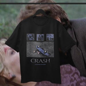 Könnte beinhalten: Ein schwarzes T-Shirt mit kurzen Ärmeln und einem Grafikdruck, der drei kleine rechteckige Bilder über einem größeren Bild eines umgekippten blauen Autos zeigt. Der Text CRASH und A FILM BY DAVID CRONENBERG ist in Weiß darunter gedruckt.