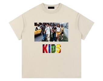 Könnte beinhalten: Ein beiges T-Shirt mit kurzen Ärmeln und einem rechteckigen Fotodruck, der eine Gruppe junger Menschen zeigt, die neben einem gelben Taxi eine Straße entlanggehen. Unter dem Bild steht das Wort KIDS in großen, bunten Blockbuchstaben gedruckt.