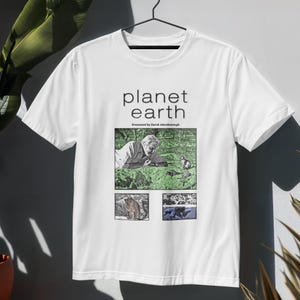 May include: A white short-sleeve t-shirt featuring the text planet earth and Presented by David Attenborough above a graphic collage. The collage includes a central image of a person lying in grass with animals, a bear, and an underwater scene.