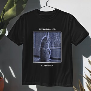 May include: A black short-sleeved t-shirt featuring a square graphic of a cat looking out a window. Above the image, the text reads THE VOID CALLED. Below the image, the text reads I ANSWERED. The cat is depicted in shades of blue and grey.