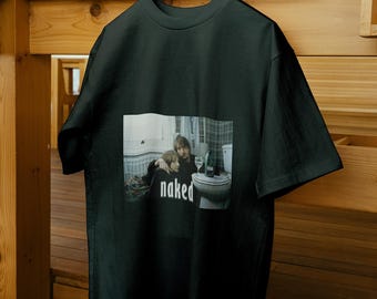 Könnte beinhalten: Ein schwarzes T-Shirt mit kurzen Ärmeln und einem rechteckigen Grafikdruck auf der Brust. Das Bild zeigt zwei Personen in einem Badezimmer mit einer Toilette und einem Weinglas, wobei das Wort "naked" in weißen Kleinbuchstaben darunter steht.