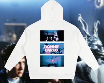 Könnte beinhalten: Ein weißer Kapuzenpullover mit einem Grafikdruck auf der Rückseite. Der Druck besteht aus drei übereinander angeordneten rechteckigen Feldern, die Szenen aus dem Film Donnie Darko zeigen, mit dem Titel Donnie Darko in rosa Buchstaben.