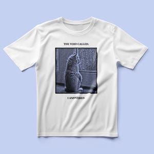 Puede incluir: Una camiseta blanca de manga corta con un gráfico cuadrado de un gatito mirando la lluvia a través de una ventana. El texto sobre el gráfico dice THE VOID CALLED. y el texto debajo dice I ANSWERED. El estampado es en tonos azules.