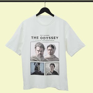 May include: A white short-sleeve t-shirt featuring a graphic print with the text "DEFY THE GODS THE ODYSSEY A FILM BY CHRISTOPHER NOLAN" above three panels showing a bearded man, a person in a head covering, a helmeted figure, and a young person.